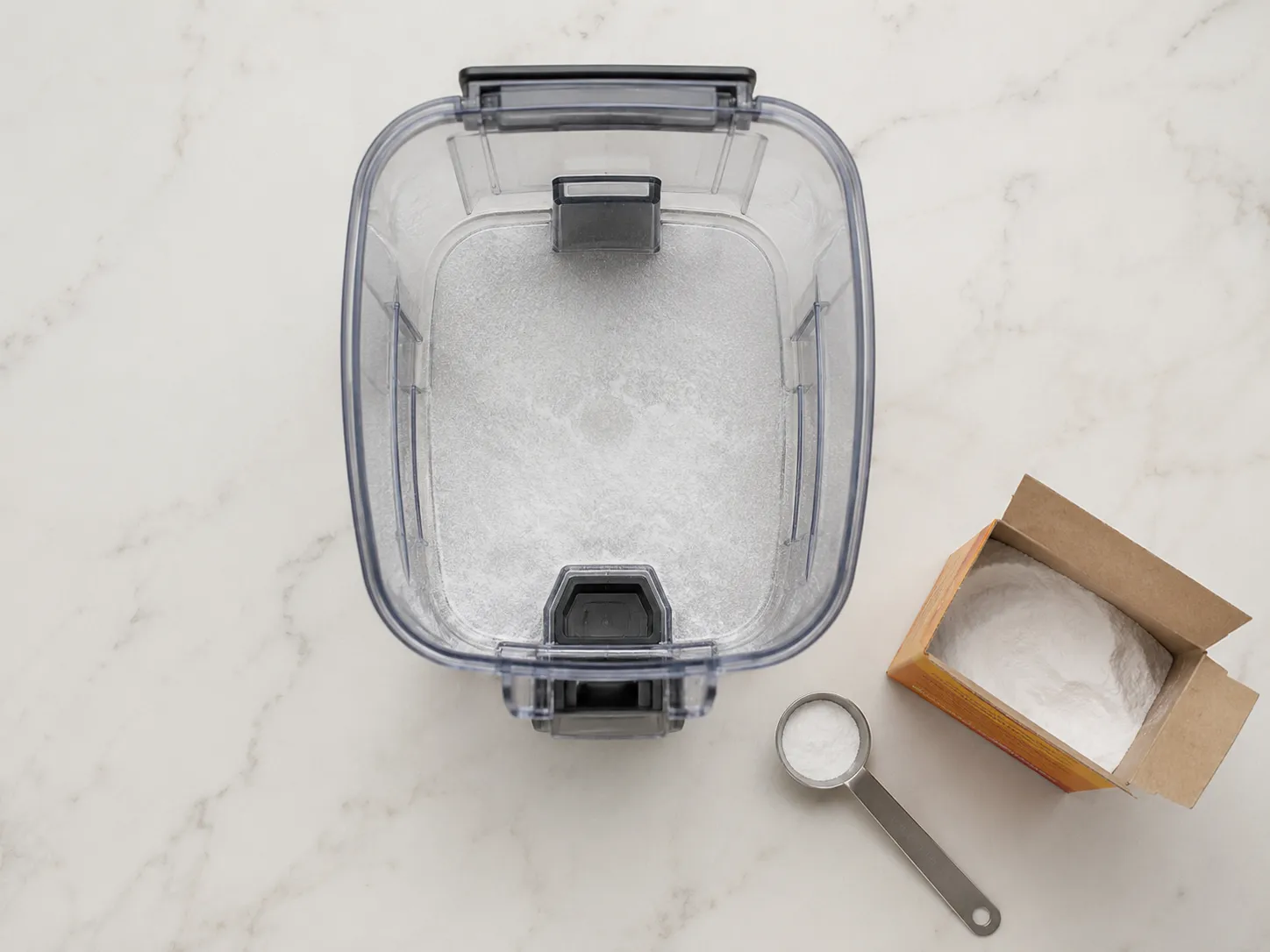 Empty transparent vacuum dustbin with baking soda sprinkled inside to remove wet-dog smell, placed on a kitchen counter with an open baking soda box and measuring spoon.