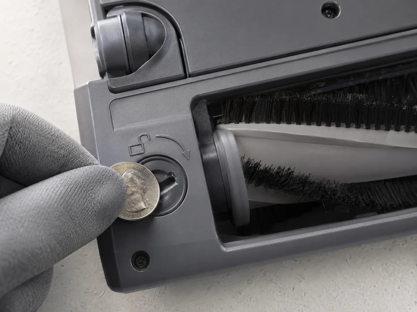 Close-up of a Bissell Featherweight vacuum brush roll lock being turned with a quarter, showing the seated roll not spinning because the lock is not yet fully unlocked.