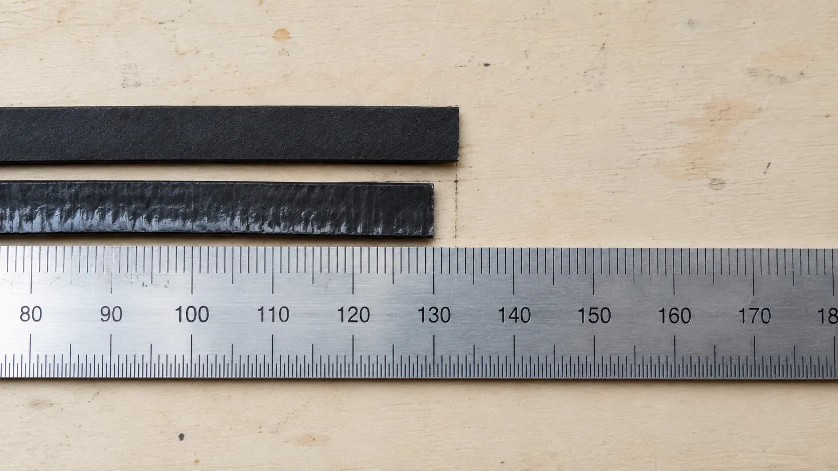 Overhead close-up of a new Bissell Style 7 vacuum belt beside a stretched used belt aligned on a steel ruler to show wear over time.