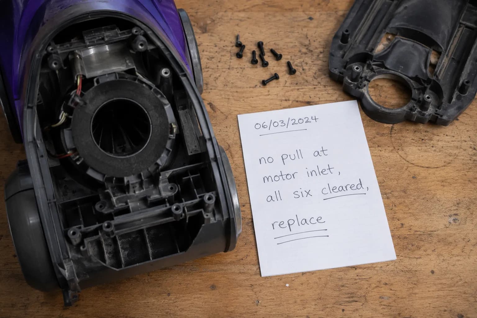 Stripped upright vacuum on a workbench beside a dated handwritten note stating there is no suction at the motor inlet after all six airflow points were cleared.