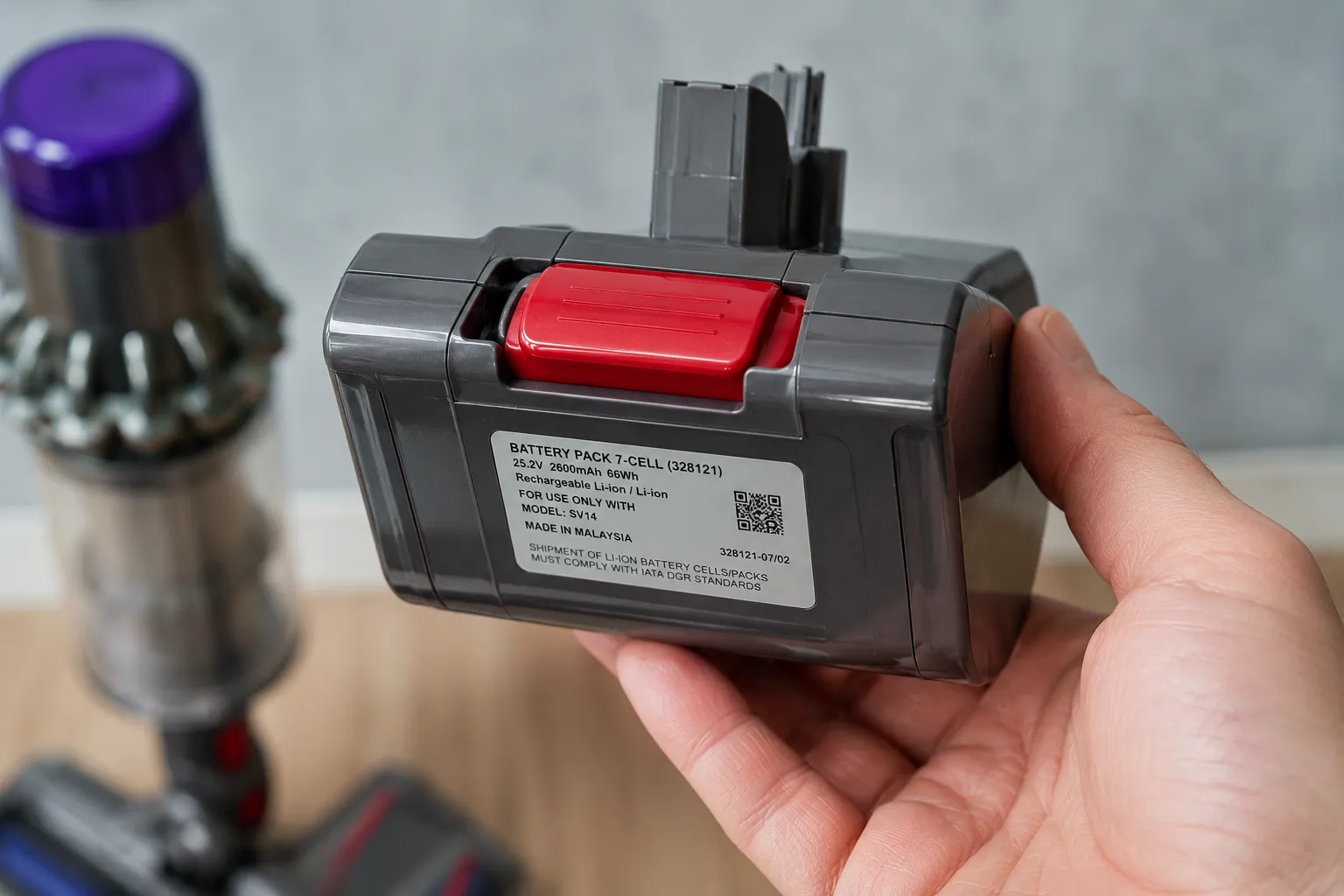 Hand holding a cordless vacuum battery pack angled to show the red click-in release button and side specification label, with the vacuum body blurred in the background.