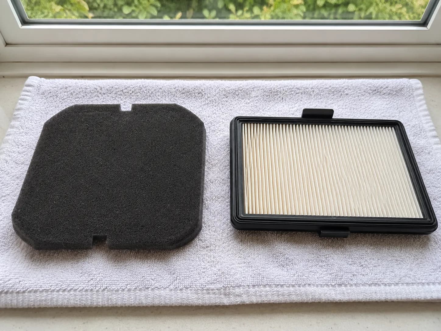 Dyson V8 foam pre-filter and pleated post-filter laid side by side on a light towel to dry near a window.