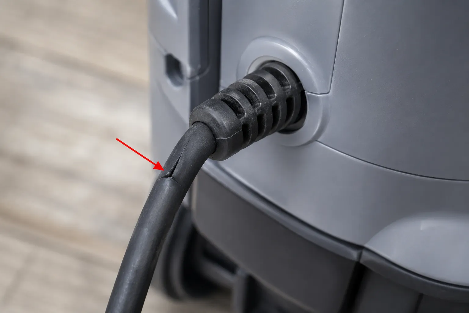 Close-up of a cracked vacuum power cord where it enters the machine body, with a red arrow marking the damaged flex point near the strain-relief collar.