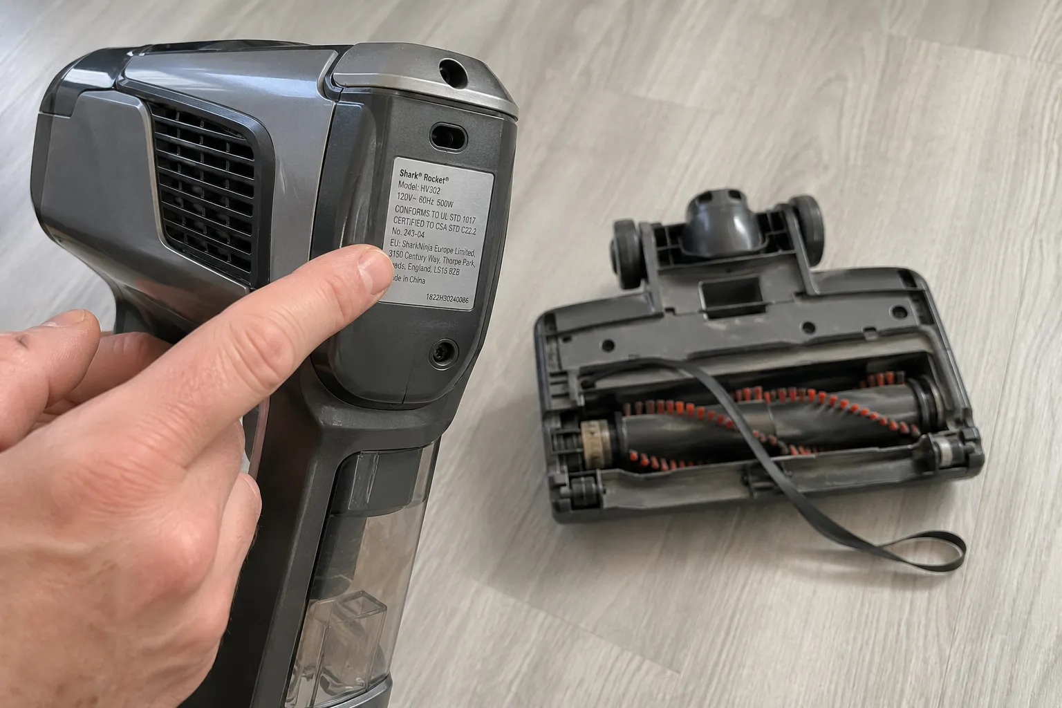 Hand pointing to the silver model-number sticker on a Shark-style Rocket handheld vacuum, with the floor nozzle brushroll and lifted belt visible nearby for belt replacement.