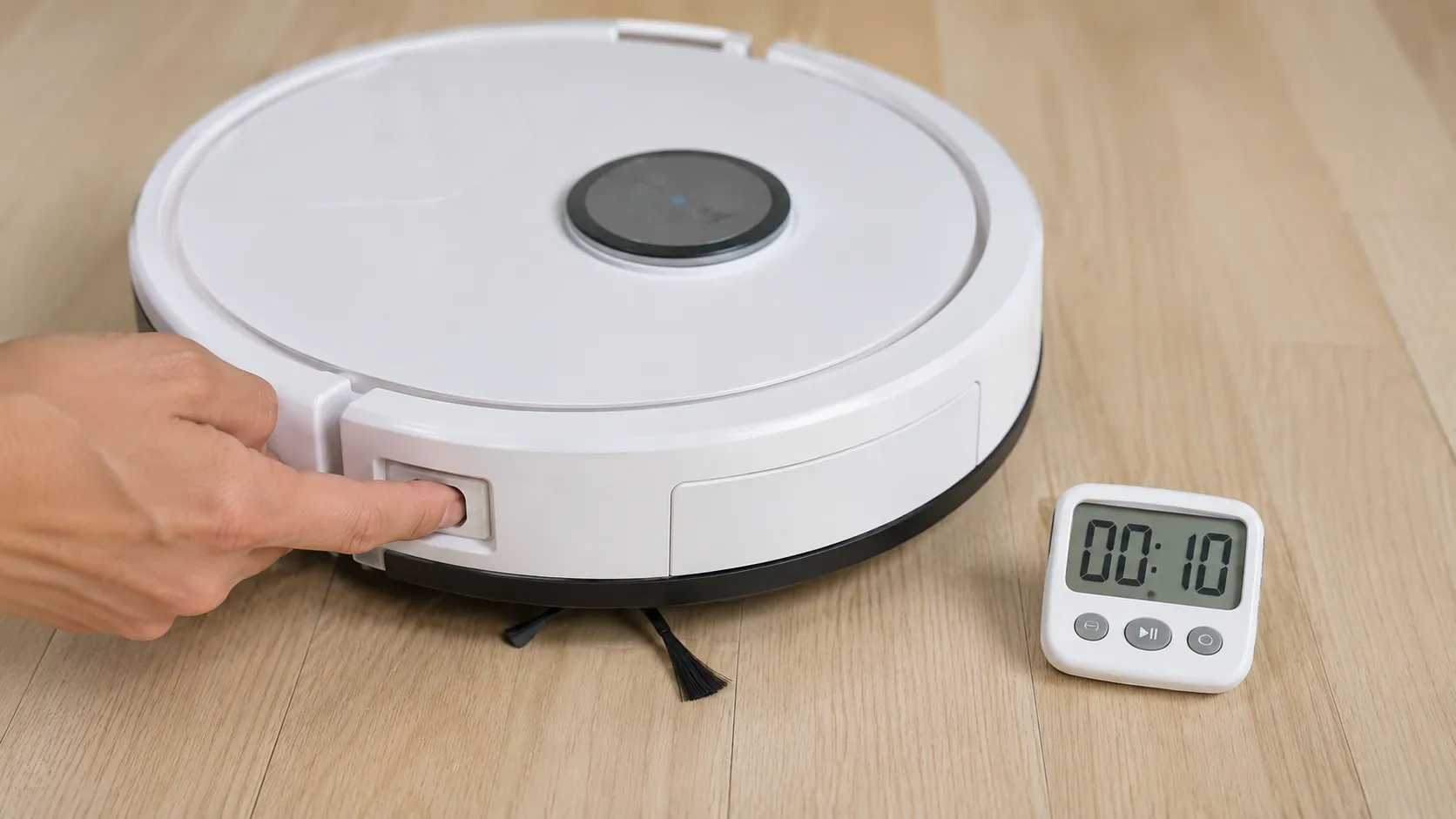 Hand pressing the side power switch on a white Shark-style robot vacuum with a stopwatch showing 10 seconds during a reset process.