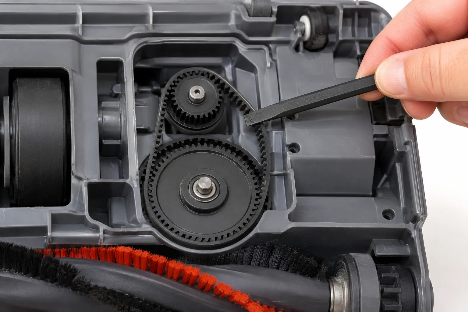 Close-up of an opened Shark-style DuoClean floor nozzle showing a hand using a plastic spudger to lift the small geared drive belt off its gear before removing the larger belt.