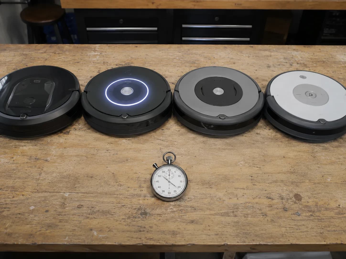 Four generations of Roomba robot vacuums lined up on a worn workbench with a stopwatch, illustrating differences in reboot timing across models