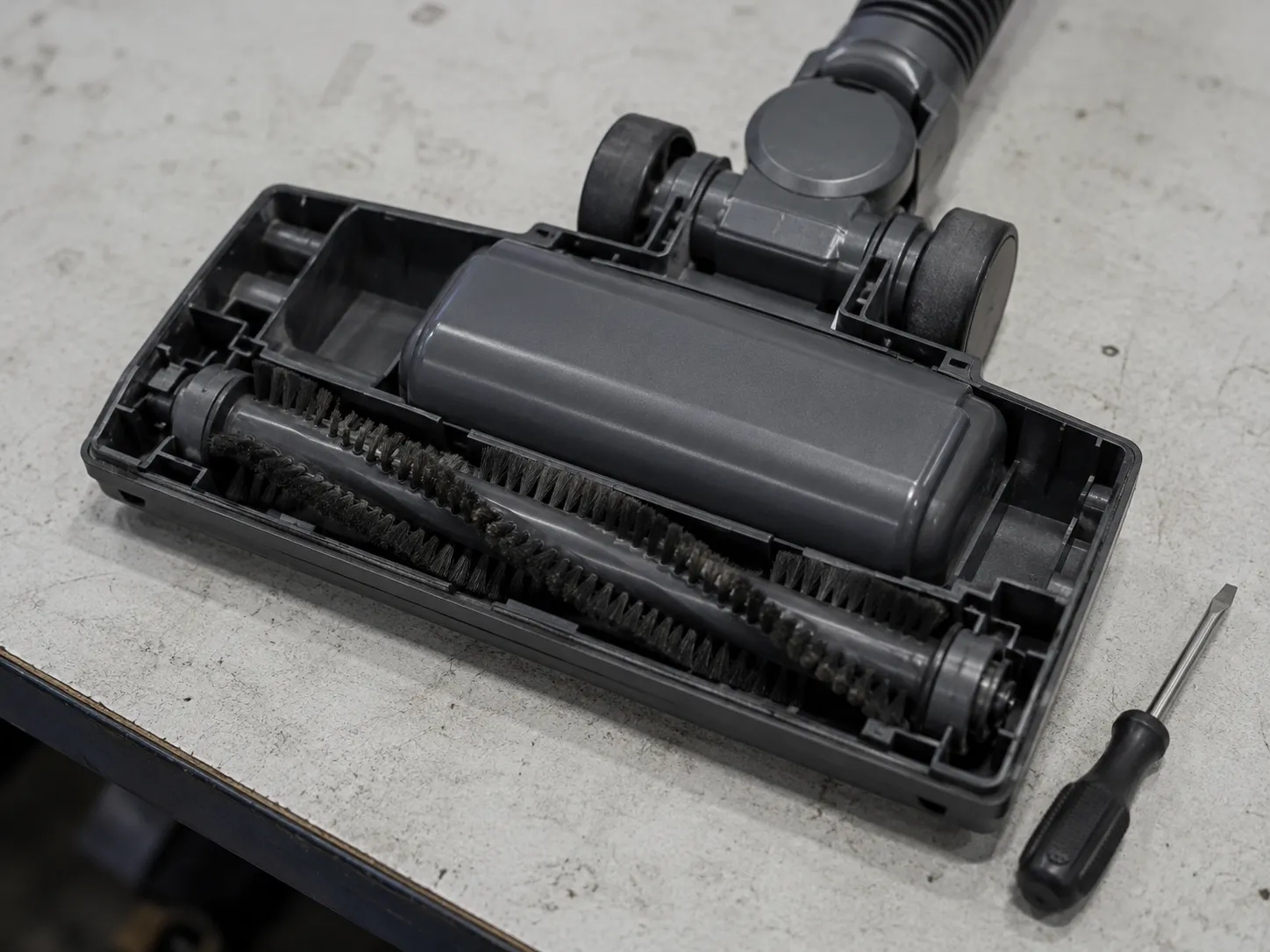 Opened direct-drive vacuum floor head showing a sealed motor housing behind the brush roll with no serviceable fasteners, beside an unused screwdriver.