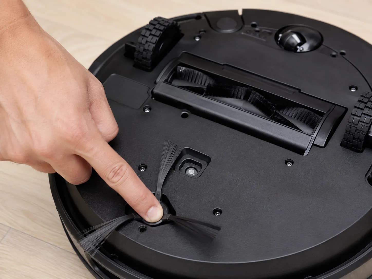 Hand pressing the recessed underside power switch on a flipped-over black Shark-style robot vacuum during a reset or power-off process.