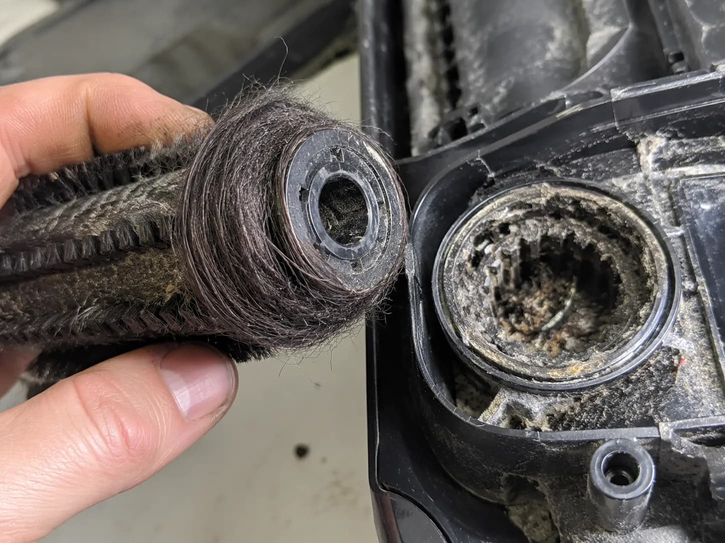 Close-up of a wet dry vacuum brush roll with tightly wound hair around the end cap beside a debris-packed drive gear socket inside the machine foot.