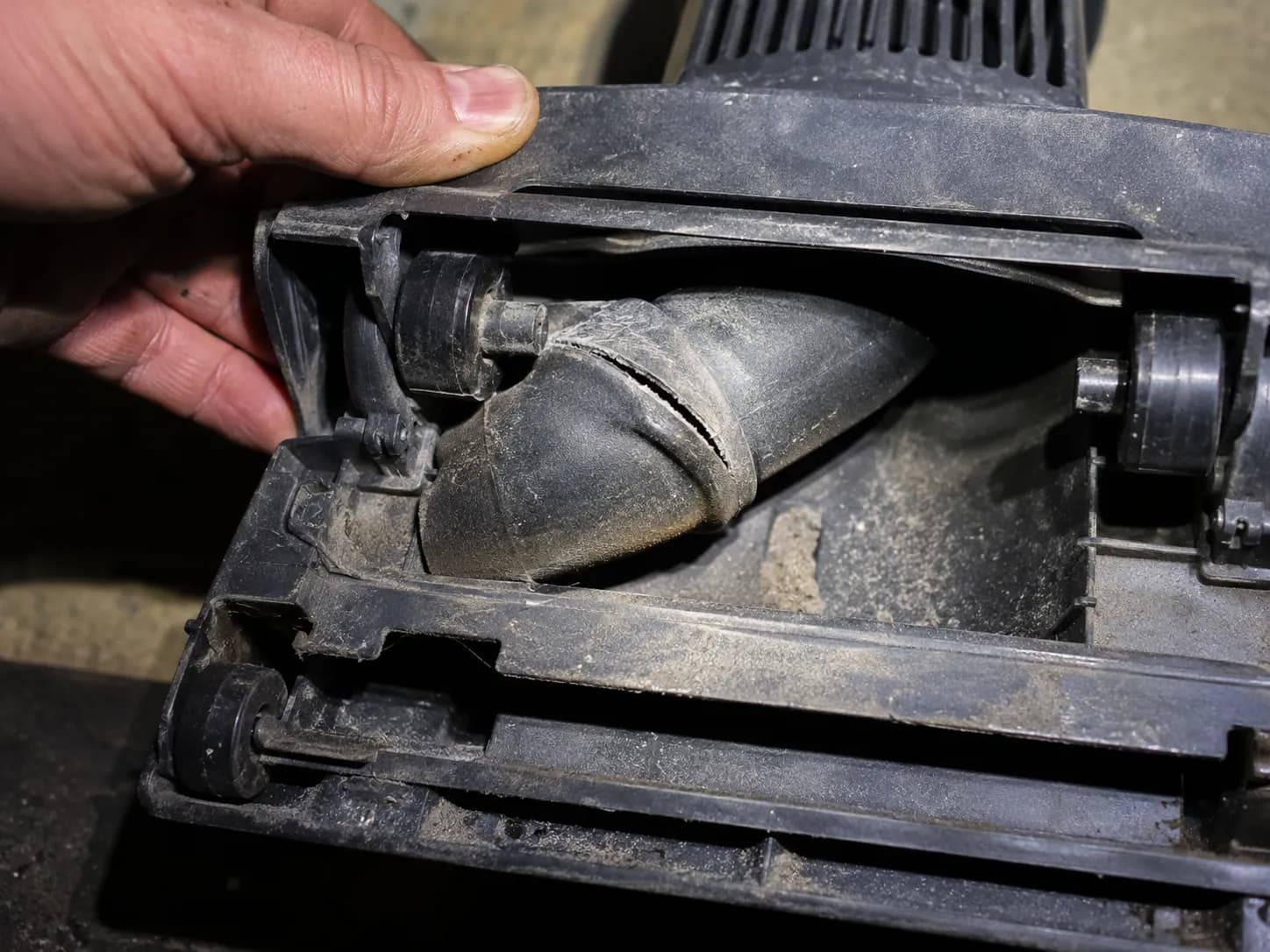 Macro repair photo inside the open floor head of a wet dry vacuum showing a short flexible rubber hose split along a crease near the pivot joint, with the brush roll removed and a hand holding the hea