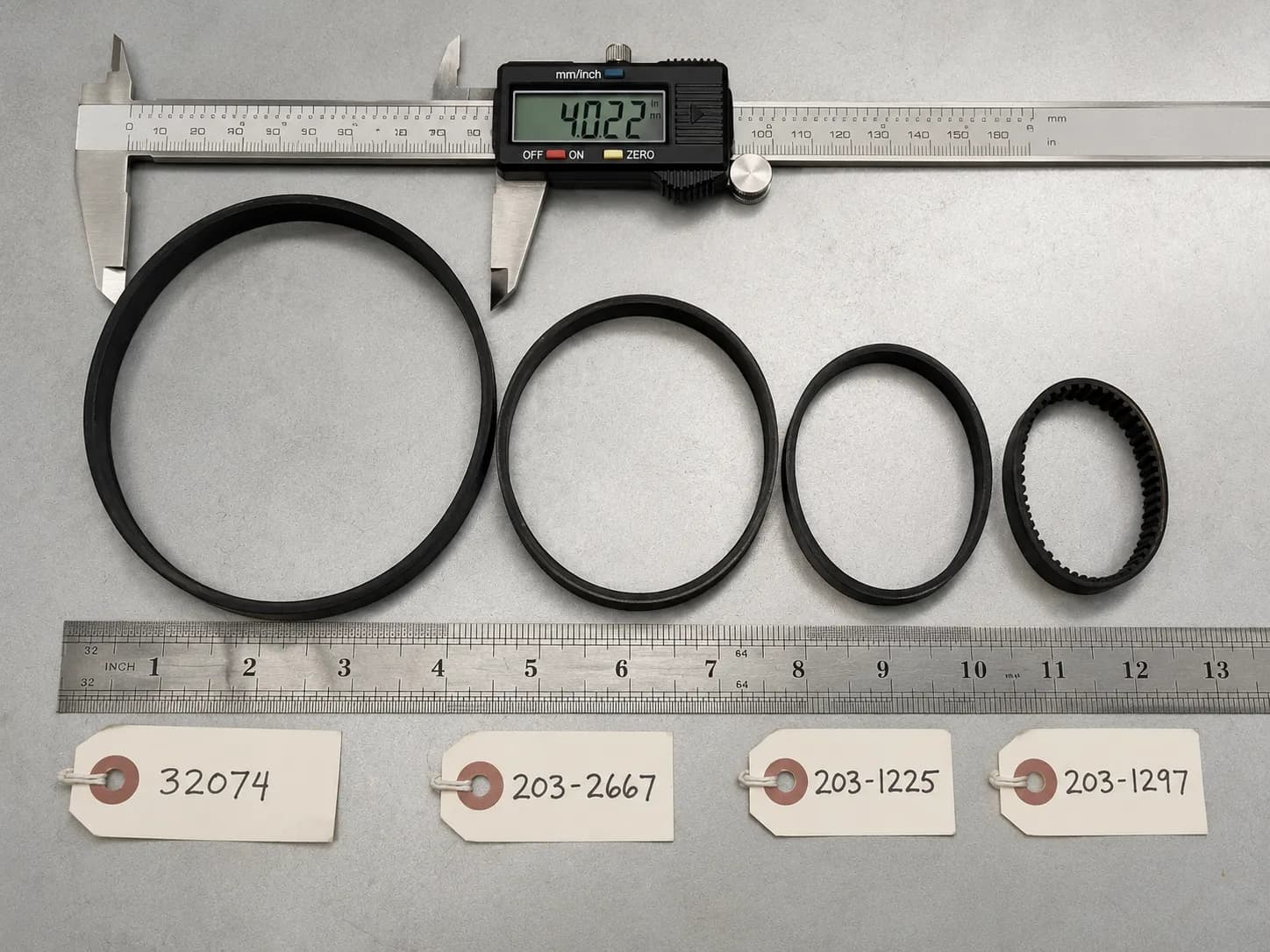 Four Bissell vacuum belts of different sizes arranged beside a steel ruler, with digital calipers measuring the largest 32074 belt.