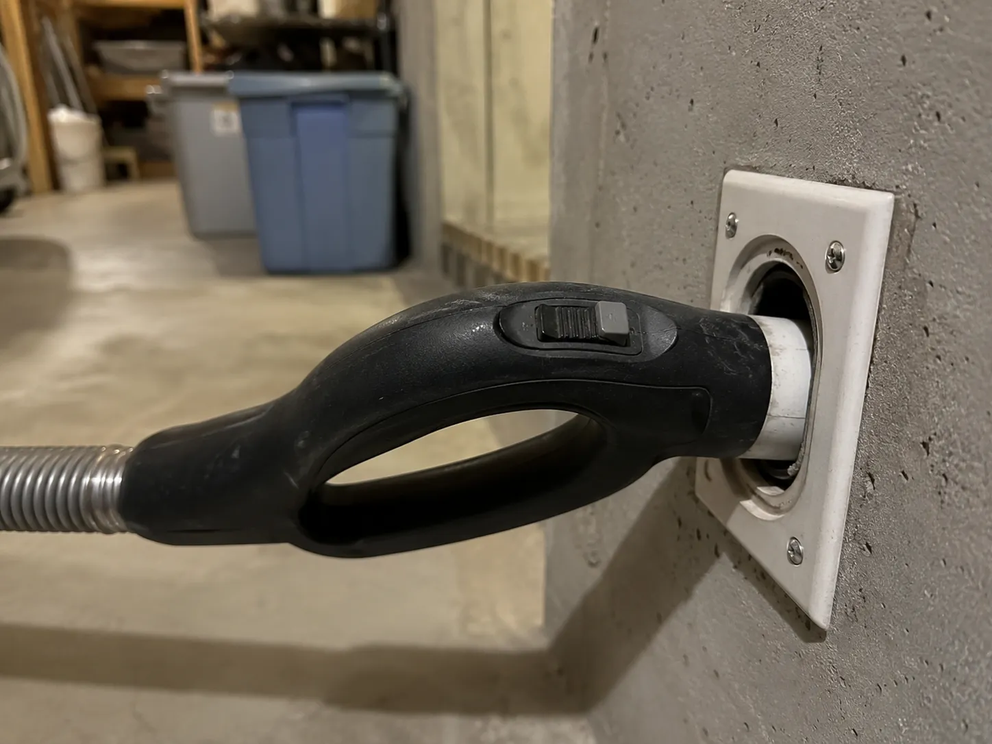 Central vacuum hose handle plugged into a basement wall inlet with the handle switch left in the on position.