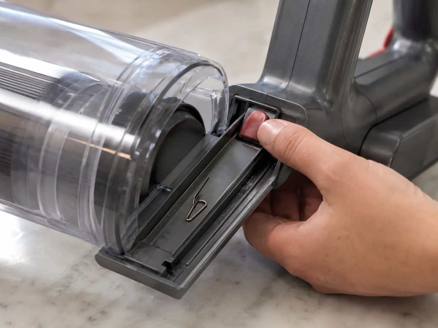 Close-up of a Dyson V7-style vacuum bin runner with a bent hairpin lodged in the track while a hand presses the red release catch during troubleshooting.