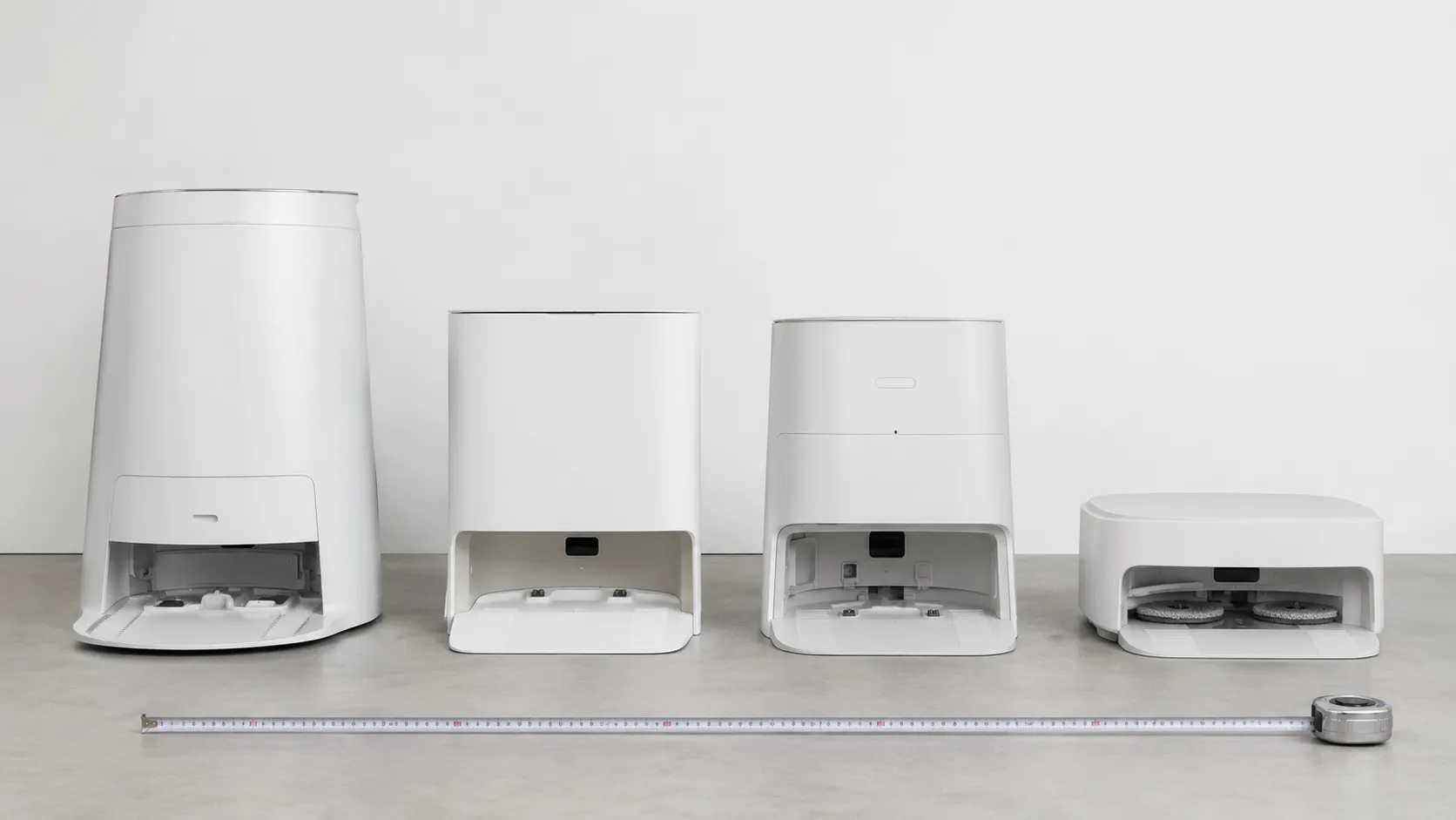 Four robot vacuum docks lined up by height, including two Clean Base models, an AutoEmpty Dock and an AutoWash Dock, with a tape measure placed in front for scale.