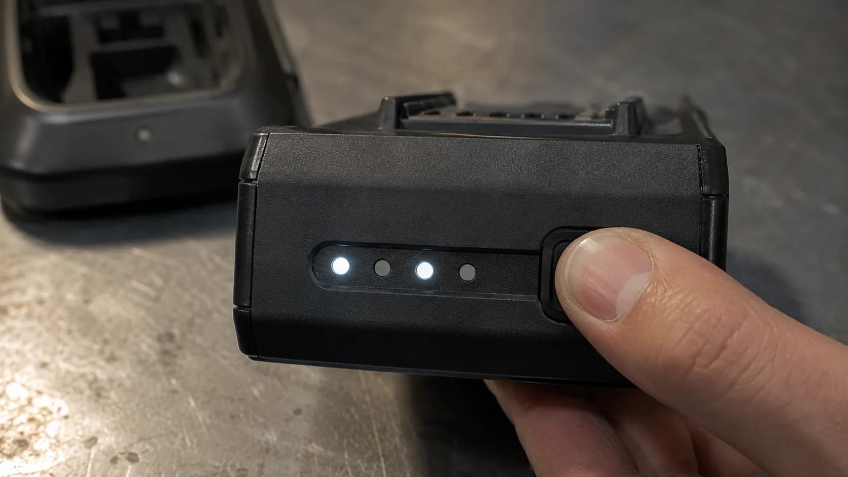 Close-up of a properly formed hand pressing the fuel-gauge button on a cordless vacuum battery pack, with the first and third LEDs lit and the charger blurred in the background.