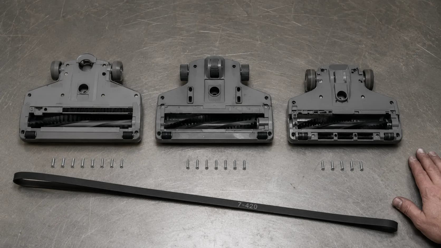 Three vacuum bottom plates arranged on a steel workbench with eight, seven and six screws lined beneath them, alongside a new Style 160 replacement belt.