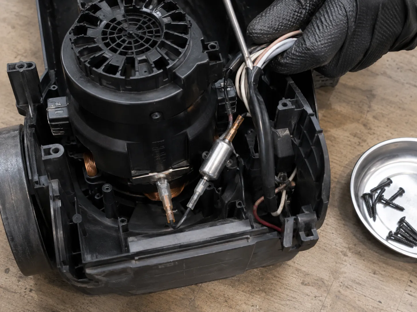 Close-up of an upright vacuum’s opened lower housing showing the thermal protector wired beside the motor while the wiring loom is held aside.
