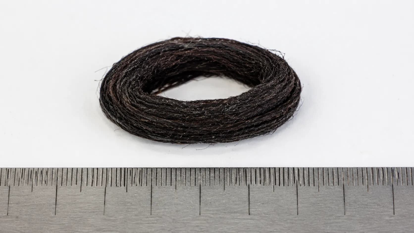Compacted 6 mm ring of dark hair and thread removed from a vacuum brushroll end cap beside a steel ruler, showing why a new belt alone did not restore performance.