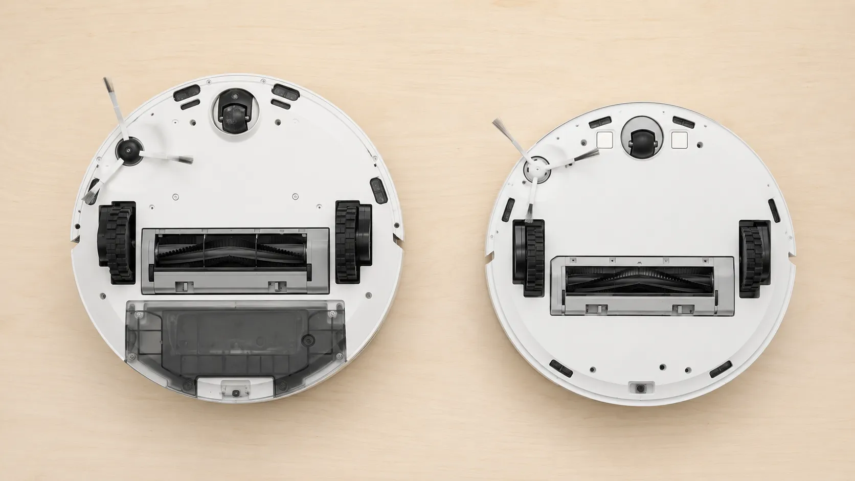 Overhead view of two white robot vacuums placed upside down side by side, comparing the underside wheels, brushes and sensor layout of an older model and a 2026 model.