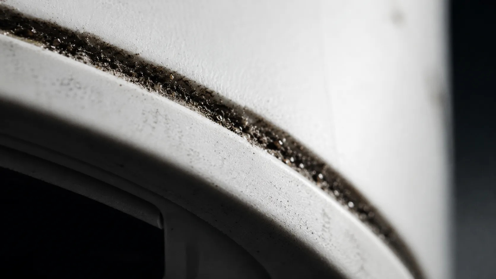 Extreme macro close-up of fine grit and grey dust packed into the narrow seam between the front bumper and main shell of a white robot vacuum.