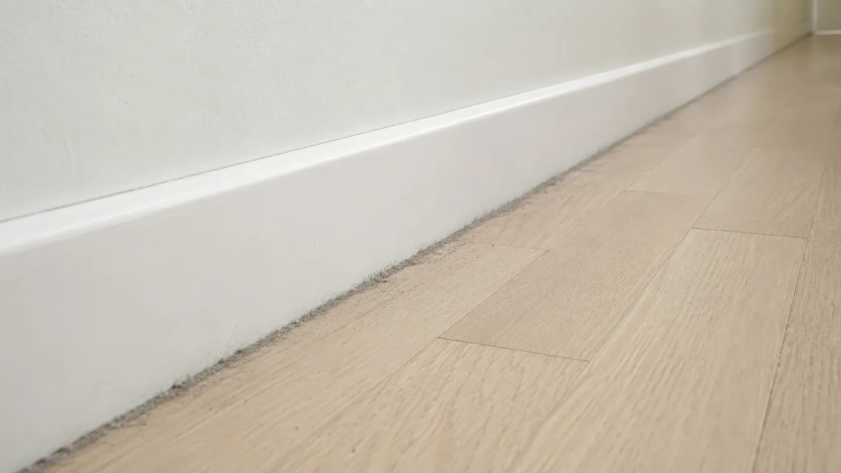 Low-angle view of a grey dust line collected along a white skirting board and pale wood floor after a robot vacuum side brush stopped working.