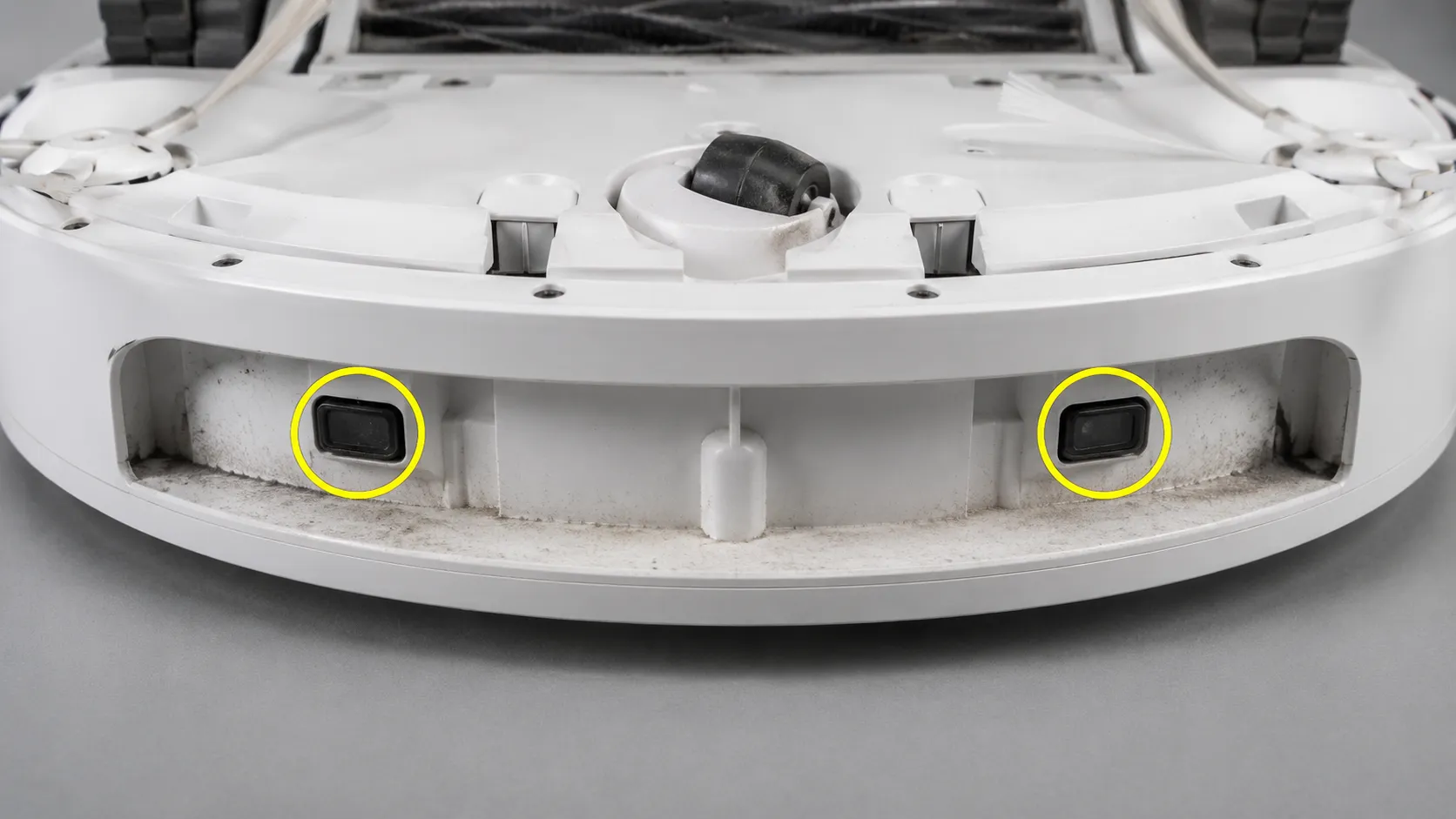 Close-up of an upside-down robot vacuum showing the hidden infrared sensor windows under the front bumper, circled in yellow with visible dust in the recess.