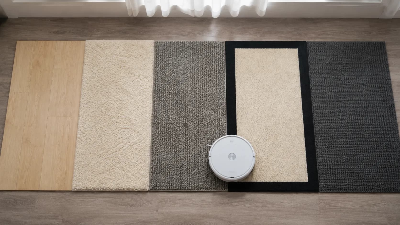 Overhead view of a Roomba 675 floor transition test across five different flooring samples, showing the robot vacuum stopping at the edge of a cream rug with a wide black border after successfully cro