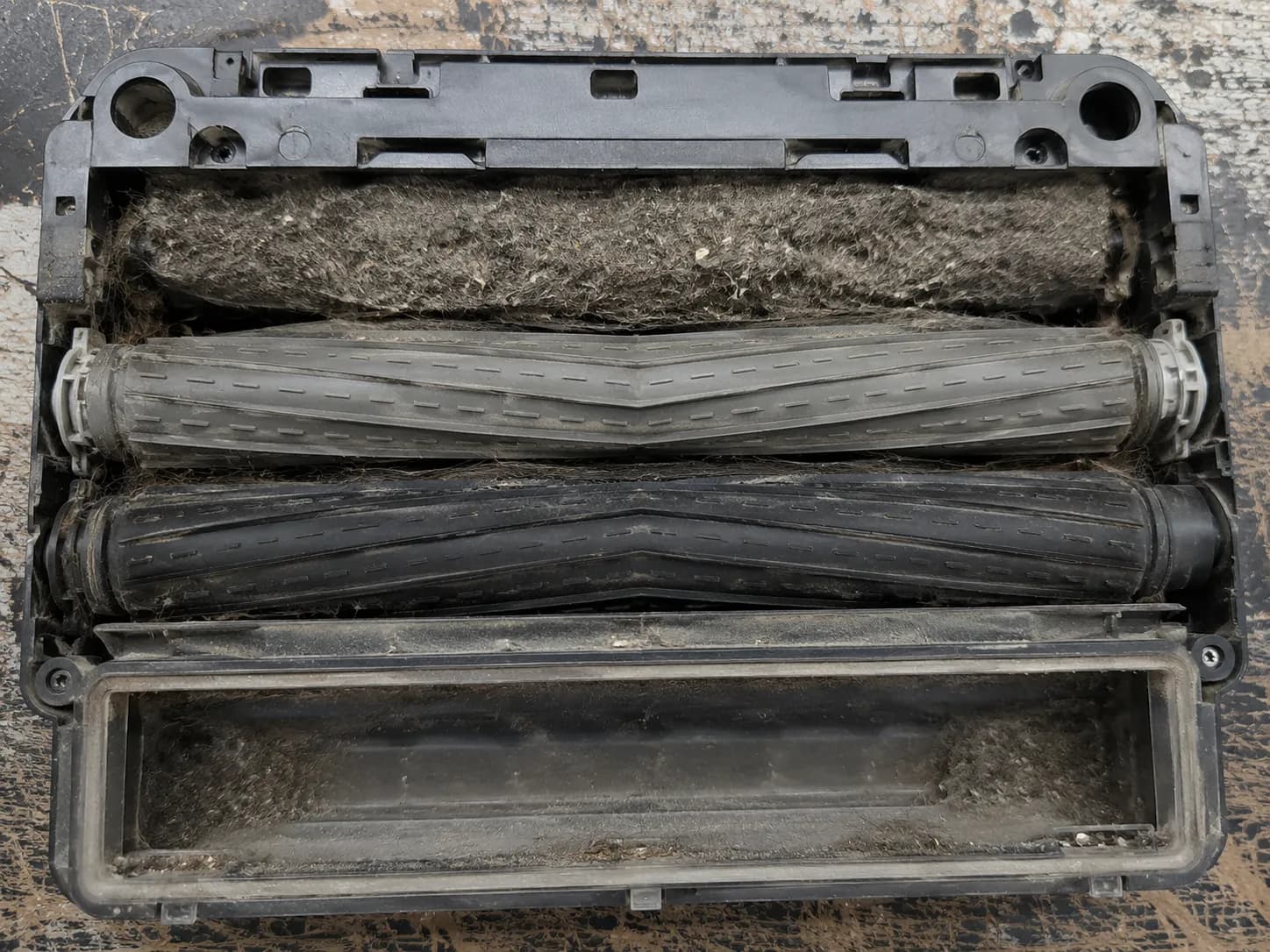 Overhead close-up of a Roomba cleaning head showing a dense clog of hair, lint and dust packed between the rubber rollers and bin opening.