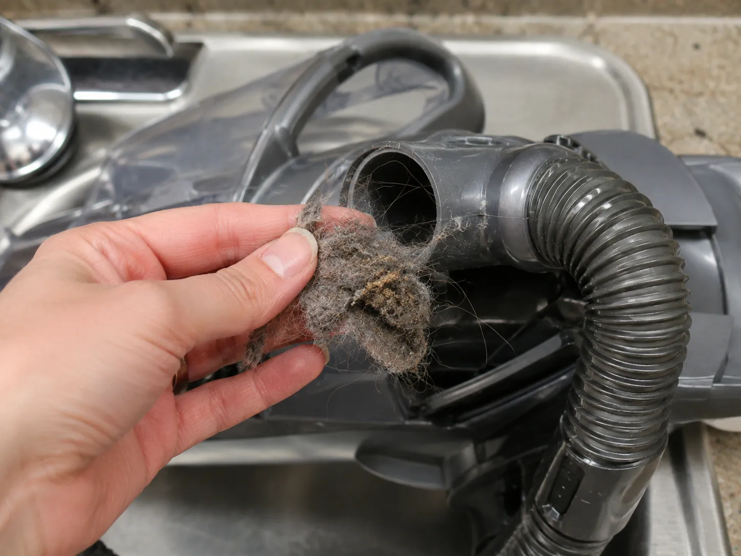 Hand removing a hair and lint clog from a Shark vacuum hose over a sink.