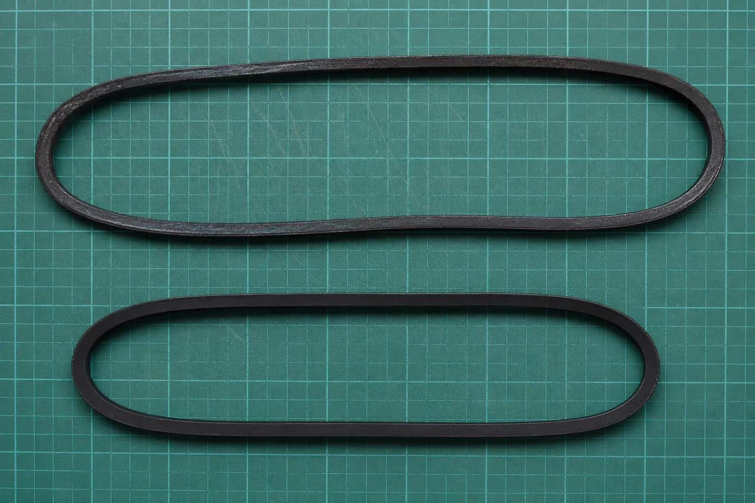 Overhead comparison of a stretched, glazed vacuum drive belt and a shorter new belt placed side by side on a green cutting mat.