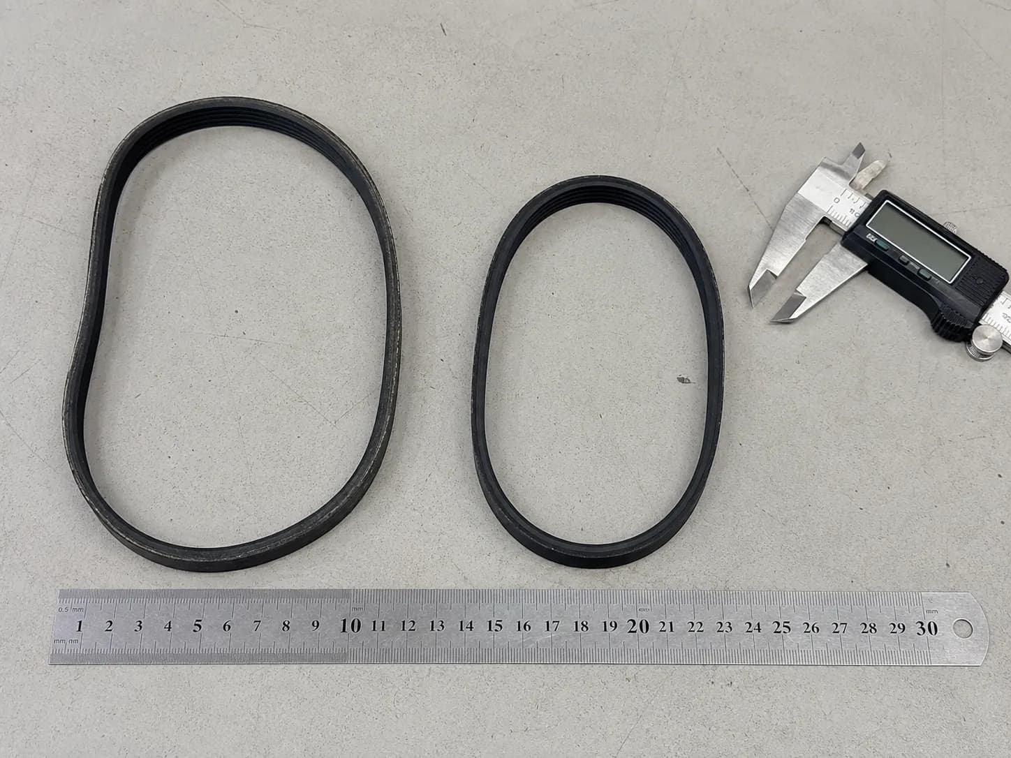 Overhead comparison of an old stretched vacuum drive belt beside a shorter new belt, with a steel ruler and digital caliper for scale.