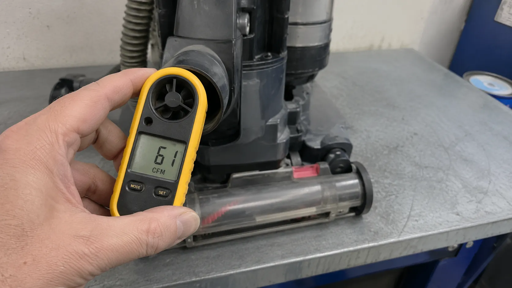 Hand holding a yellow anemometer at an upright vacuum hose cuff, displaying an airflow reading of 61 CFM during a suction test.