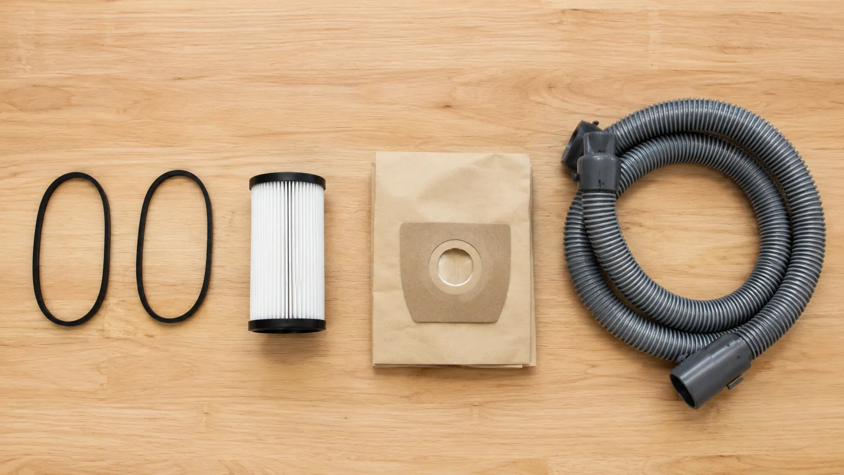 Overhead flat lay of five replacement vacuum parts on a light wood workbench, including two drive belts, a pleated filter, paper vacuum bags and a coiled hose.